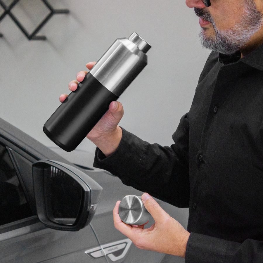 Botella térmica de acero inoxidable con cuerpo negro mate, sostenida junto a un automóvil